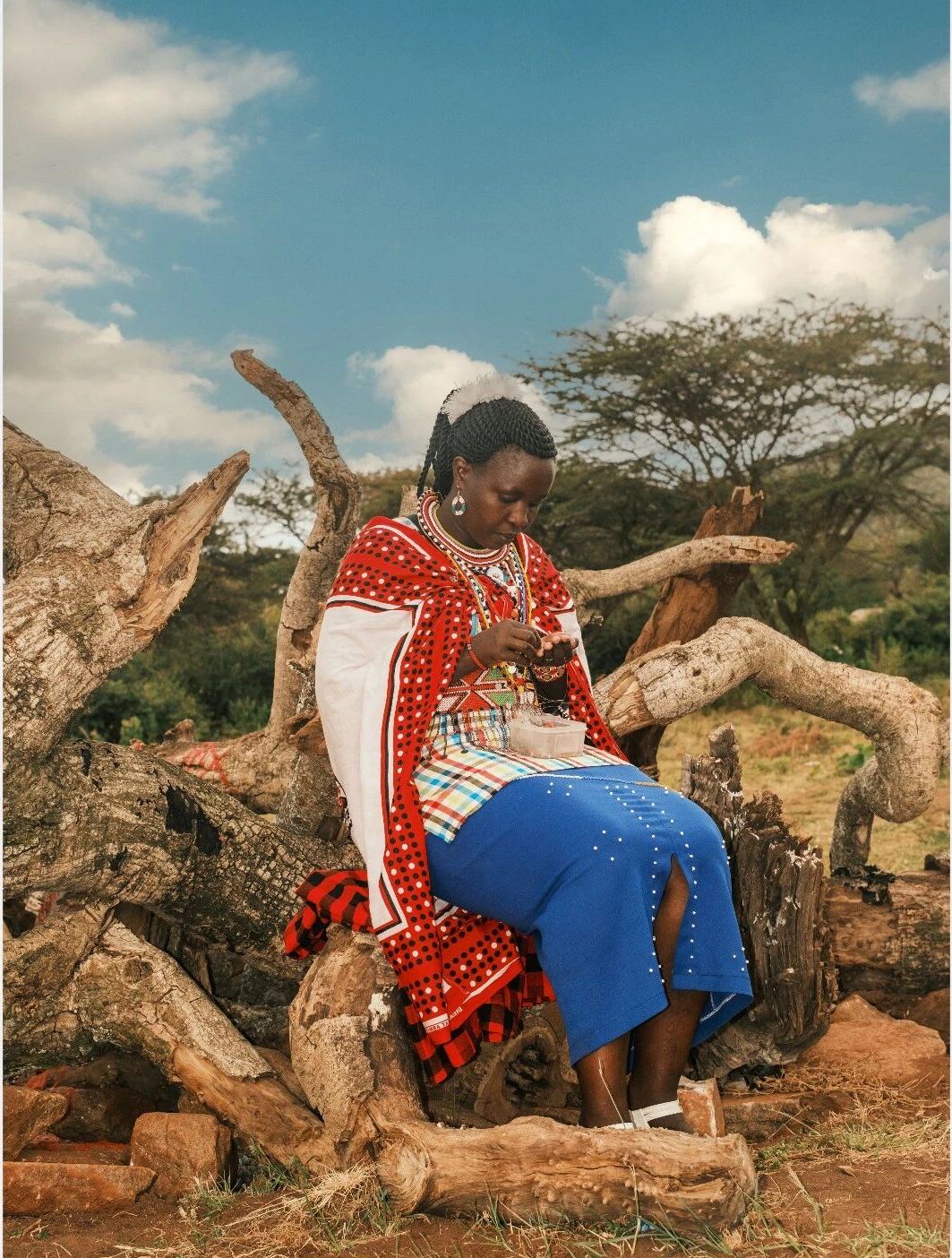 A Maasai woman in a red beaded cape threading beads while seated on a fallen tree