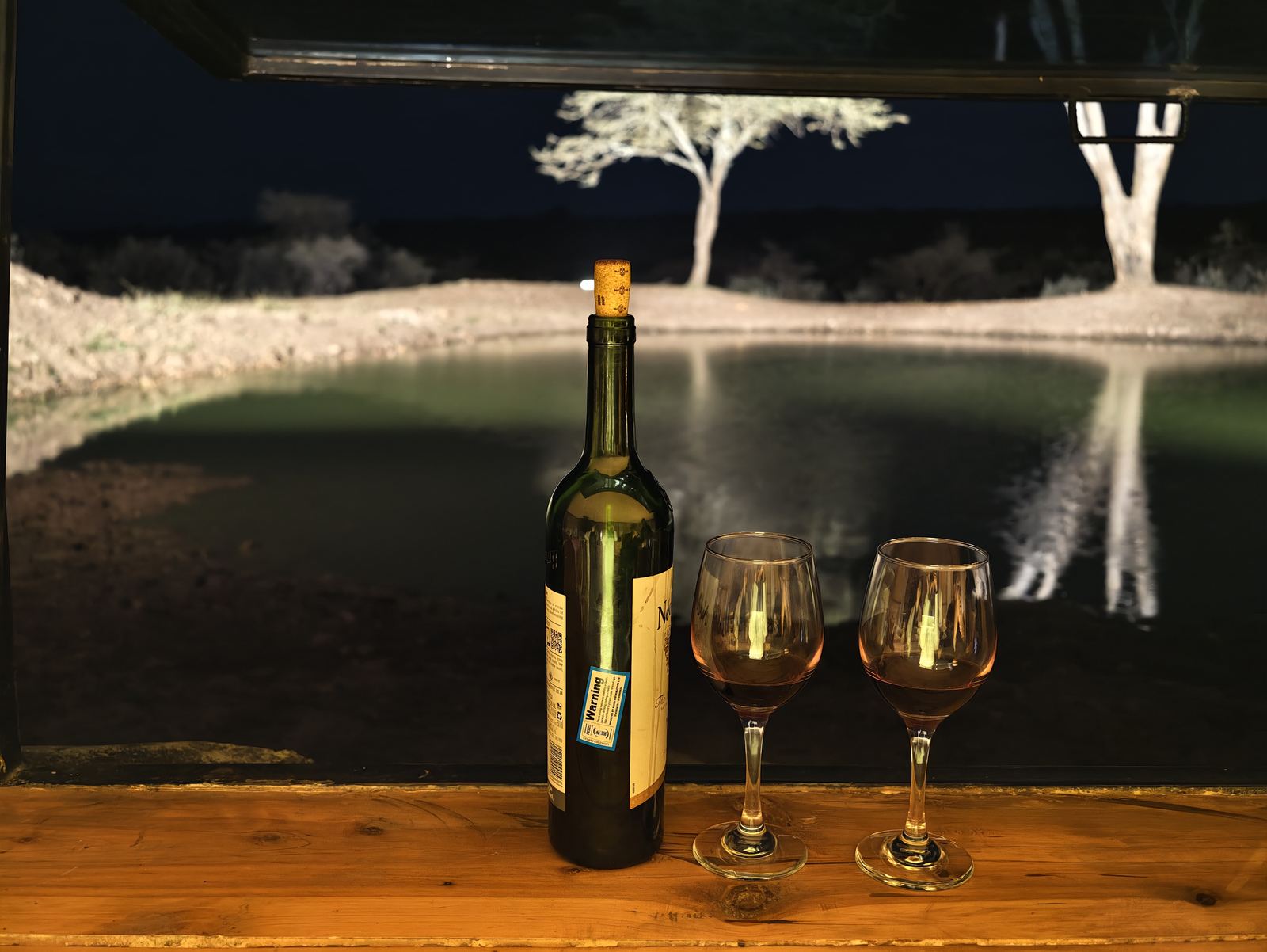 A bottle of red wine and two glasses on the hide’s ledge, looking out over a softly lit waterhole at night